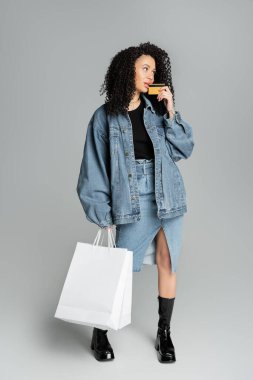 Full length of stylish woman holding credit card and shopping bags on grey background 