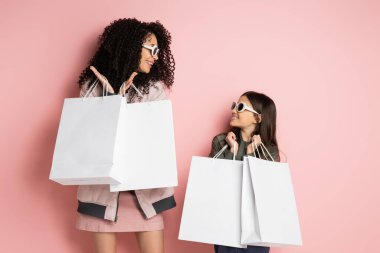 Smiling preteen girl holding shopping bags near mom in sunglasses on pink background