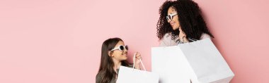 Cheerful woman and daughter in sunglasses holding shopping bags on pink background, banner 