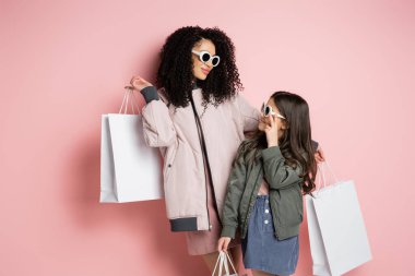 Trendy woman and kid in sunglasses holding shopping bags on pink background 