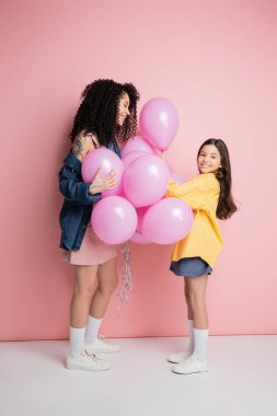 Full length of cheerful girl holding balloons near mom on pink background 