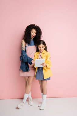 Stylish woman using digital tablet with daughter on pink background 