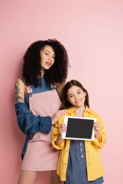 Stylish woman hugging kid holding digital tablet with blank screen on pink background 