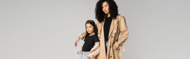 Stylish mom and kid in trench coats posing and looking at camera isolated on grey, banner 