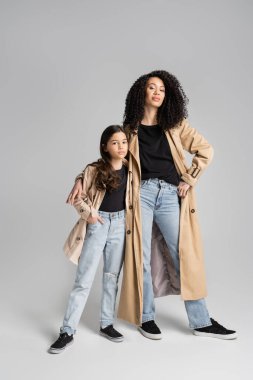 Trendy woman and girl in trench coats looking at camera on grey background 