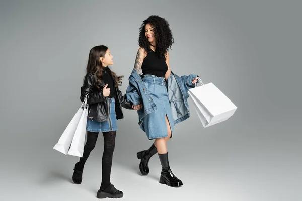 Trendy mother and daughter holding shopping bags while walking on grey background