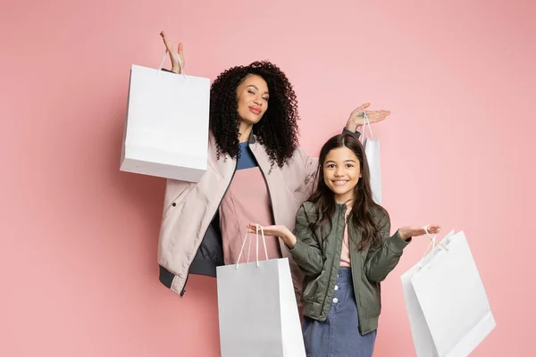 Fashionable mom and daughter holding shopping bags and looking at camera on pink background