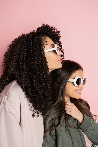 Curly woman in sunglasses standing near daughter in bomber jacket on pink background