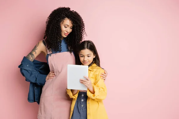 stock image Trendy woman hugging daughter with digital tablet on pink background 