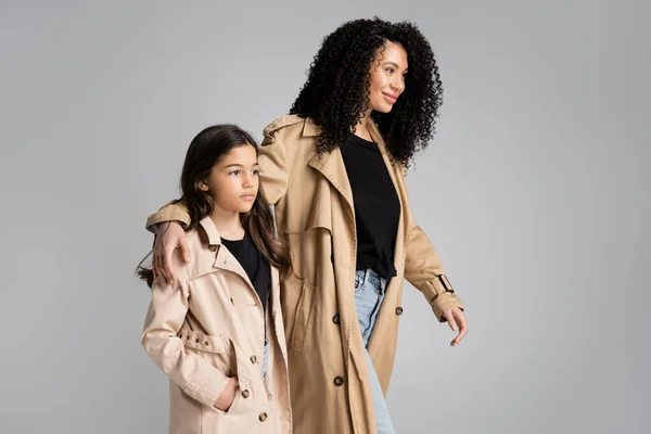 Smiling curly woman in trench coat walking near daughter isolated on grey