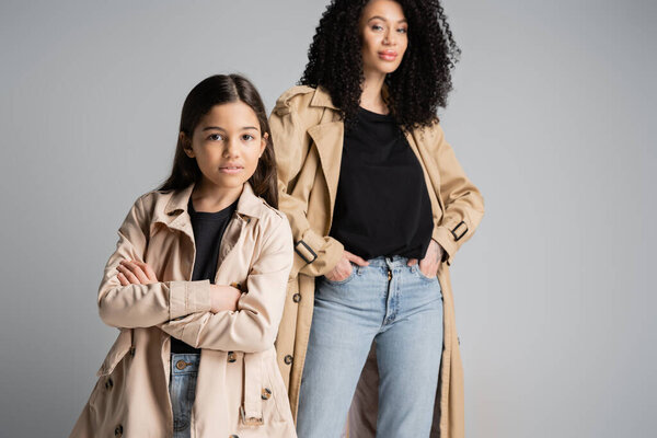 Preteen kid in trench coat crossing arms near blurred mom isolated on grey 