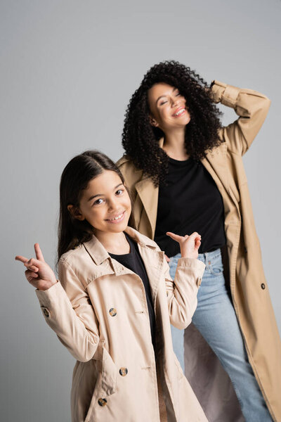 Smiling girl in trench coat showing peace sign near mom isolated on grey 
