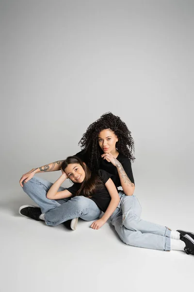 stock image Tattooed woman and daughter in jeans and t-shirts smiling at camera on grey background 