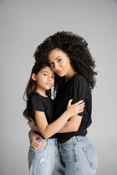 Stylish mother and daughter in jeans and t-shirts looking at camera isolated on grey