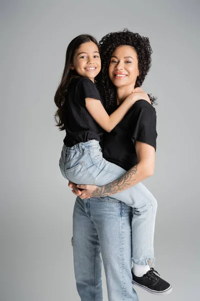 Tattooed mother lifting daughter in jeans and t-shirt isolated on grey