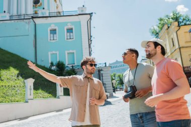 african american tourist with vintage camera looking away near bearded man and tour guide pointing with hand on Andrews descent in Kyiv clipart