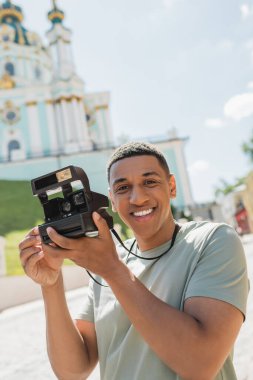 Kyiv 'deki bulanık St. Andrews Kilisesi yakınlarında gülümseyen klasik kameralı kaygısız Afrikalı Amerikalı turist