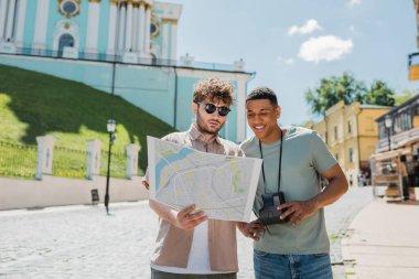 Güneş gözlüklü bir tur rehberi ve klasik kameralı Afro-Amerikan gezgin Ukrayna 'nın başkenti Kyiv' de şehir haritasına bakıyor.