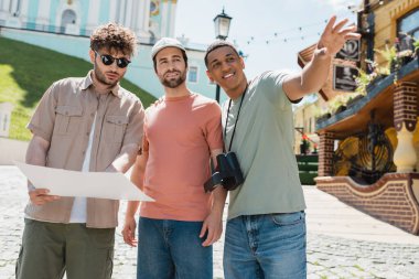 happy african american tourist with vintage camera looking away and pointing with hand near friend and tour guide with map in Kyiv clipart