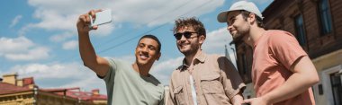 happy african american traveler taking selfie with tour guide in sunglasses and bearded man on Andrews descent in Kyiv, banner clipart