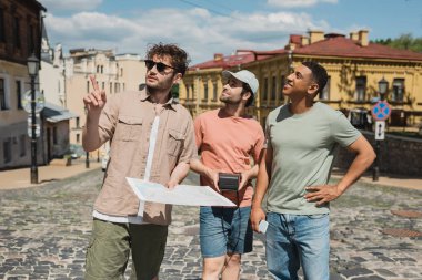  tour guide in sunglasses holding map and pointing with finger near interracial travelers on Andrews descent in Kyiv clipart