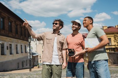 happy tour guide in sunglasses pointing with finger near interracial tourists with city map and vintage camera on Andrews descent in Kyiv clipart