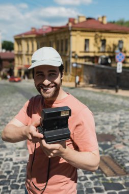 cheerful bearded traveler in sun hat holding vintage camera on Andrews descent in Kyiv, Ukraine clipart