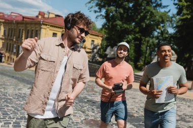 positive guide in sunglasses and headset leading tour for multiethnic tourists with map and vintage camera on Andrews descent in Kyiv clipart