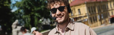 portrait of carefree tour guide in sunglasses and headset smiling at camera on blurred Andrews descent in Kyiv, banner clipart
