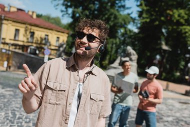 young and carefree guide in sunglasses and headset pointing with finger near blurred interracial tourists on Podil district in Kyiv clipart