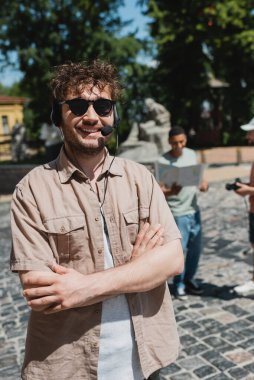 Güneş gözlüklü ve kulaklıklı genç ve mutlu tur rehberi yaz günü Kyiv 'de Andrews' e doğru inerken çapraz kollarıyla ayakta duruyor.