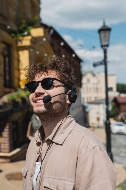 young and curly tour guide in sunglasses and headset smiling on blurred Andrews descent in Kyiv clipart
