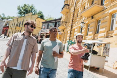 bearded tourist in sun cap holding vintage camera and pointing with hand during excursion on Podil district in Kyiv  clipart