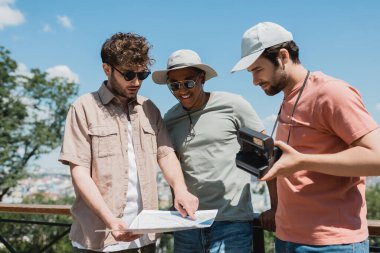 young tour guide in sunglasses looking at travel map near interracial tourists with vintage camera in city park clipart