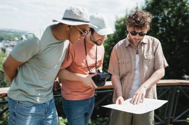 Güneş gözlüklü genç tur rehberi şehir parkındaki gezi sırasında güneş şapkalı ırklar arası turistlerin haritasını gösteriyor.