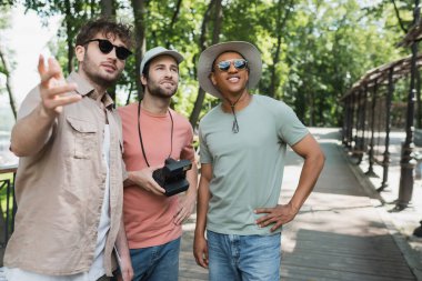 interracial travelers with vintage camera smiling and looking away near tour guide pointing with hand in city park clipart