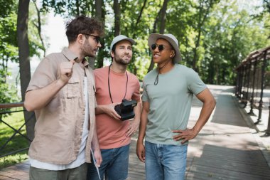 bearded tourist with vintage camera looking away near multiethnic men during excursion in summer park clipart