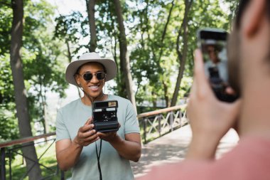 blurred man with smartphone taking photo of carefree african american tourist in sunglasses posing with vintage camera in park clipart