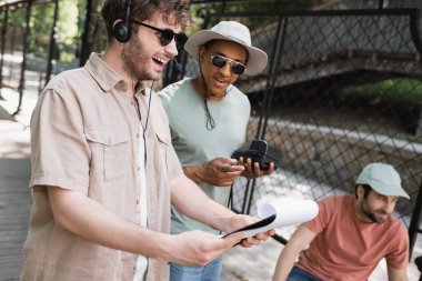 excited tour guide in headset looking at route on clipboard near multiethnic tourists in sun hats on city street clipart