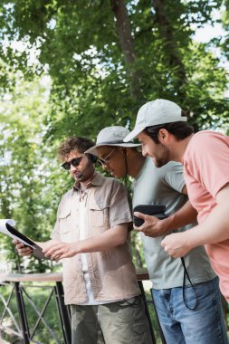 young guide in sunglasses and headset holding map near interracial travelers wearing sun hats in city park clipart