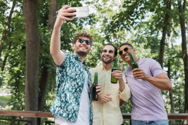 Mutlu ve son moda güneş gözlüklü adam yeşil parkta bira içen ırklar arası arkadaşlarla akıllı telefondan selfie çekiyor.