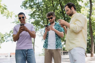 Şehir parkında akıllı telefonlar kullanarak şık yaz kıyafetleri ve güneş gözlüğü takan çok kültürlü adamlar.