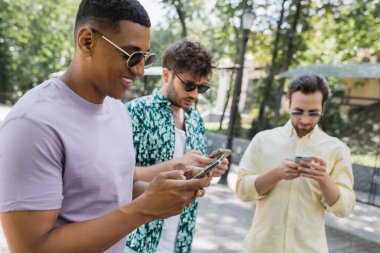 Mutlu ve şık Afrikalı Amerikalı adam şehir parkında bulanık arkadaşlarının yanında akıllı telefon kullanıyor.