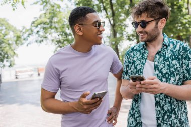 Güneş gözlüklü neşeli Afrikalı Amerikalı adam cep telefonu tutuyor ve şehir parkında trend arkadaşlarıyla konuşuyor.