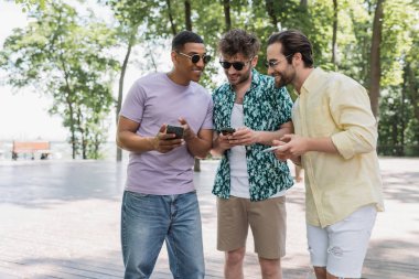 Neşeli Afrikalı Amerikalı adam akıllı telefon kullanıyor ve parkta güneş gözlüklü arkadaşlarıyla konuşuyor. 