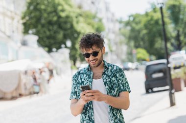 Şehir sokağında güneş gözlüklü, yürüyen ve akıllı telefon kullanan çok neşeli bir adam. 