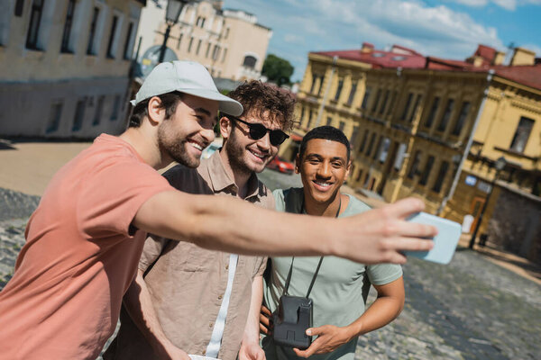 happy tourist in sun cap taking selfie with multiethnic men during excursion on Andrews descent in Kyiv