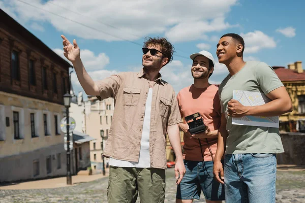 stock image tour guide in sunglasses pointing with hand near multiethnic men with map and vintage camera on Andrews descent in Kyiv