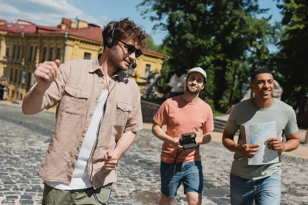stock image positive guide in sunglasses and headset leading tour for multiethnic tourists with map and vintage camera on Andrews descent in Kyiv