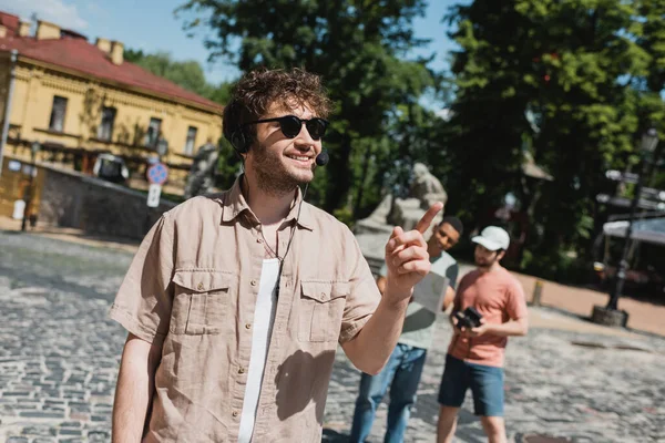 tour guide in sunglasses and headset smiling and pointing with finger near blurred multiethnic tourists on Andrews descent in Kyiv